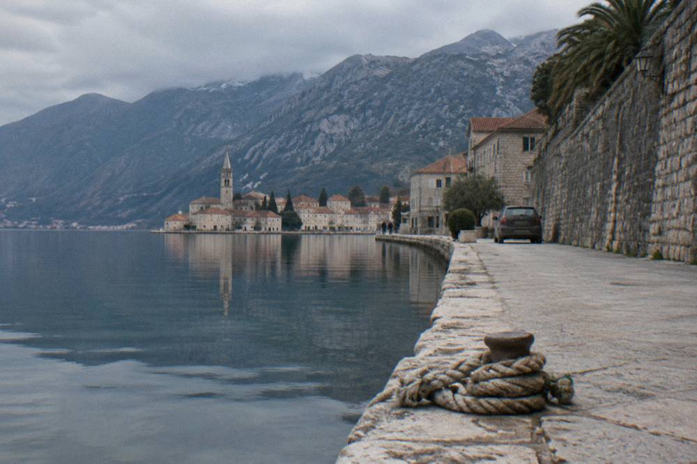 Rental car parked on a stone quay beside a walled old town and calm water