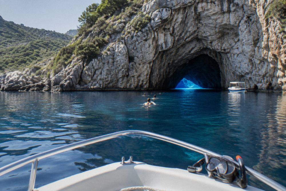 Swimmers in clear blue water beside a boat anchored at the mouth of a sea cave