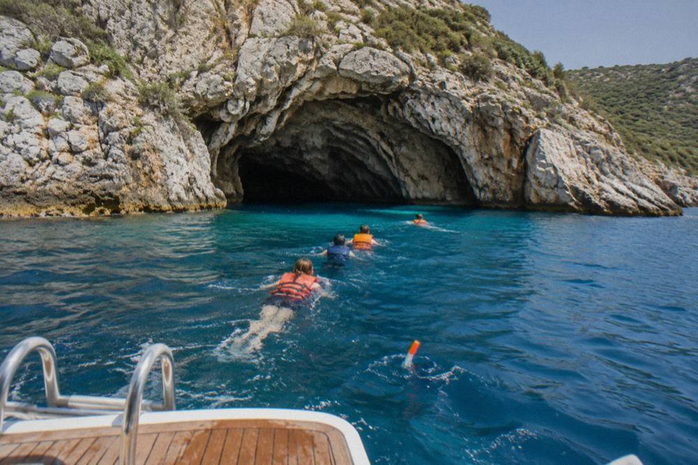 Swimmers in life jackets heading from an anchored speedboat toward a rocky cave entrance on a calm blue sea