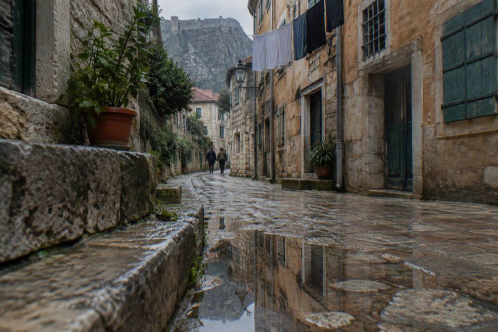 Narrow stone lane inside Kotor Old Town with old shutters and a distant glimpse of the fortress wall