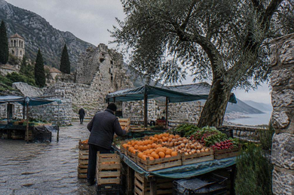 A market stall with handwritten signs and crates of fresh produce in a Montenegrin town square