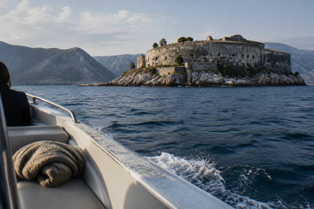 A small private speedboat cruising past a stone fortress island at the entrance to the Bay of Kotor