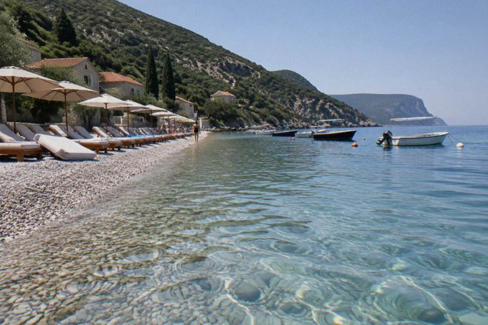 A pebble beach with sunbeds and small boats moored in clear shallow water below green hills