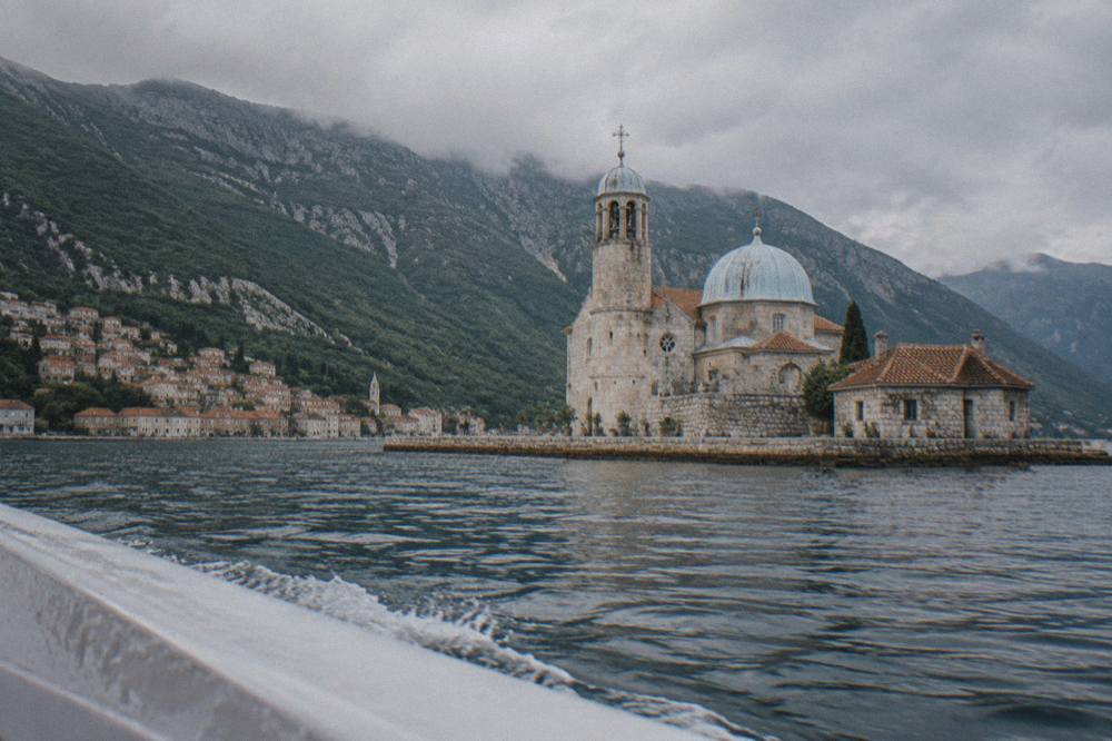 Speedboat passing a stone island church and waterfront houses along the Bay of Kotor