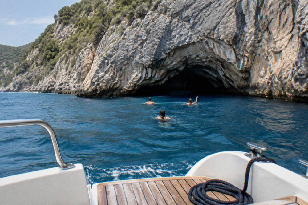 A speedboat anchored in open water near a rocky cliff with swimmers heading toward a cave entrance