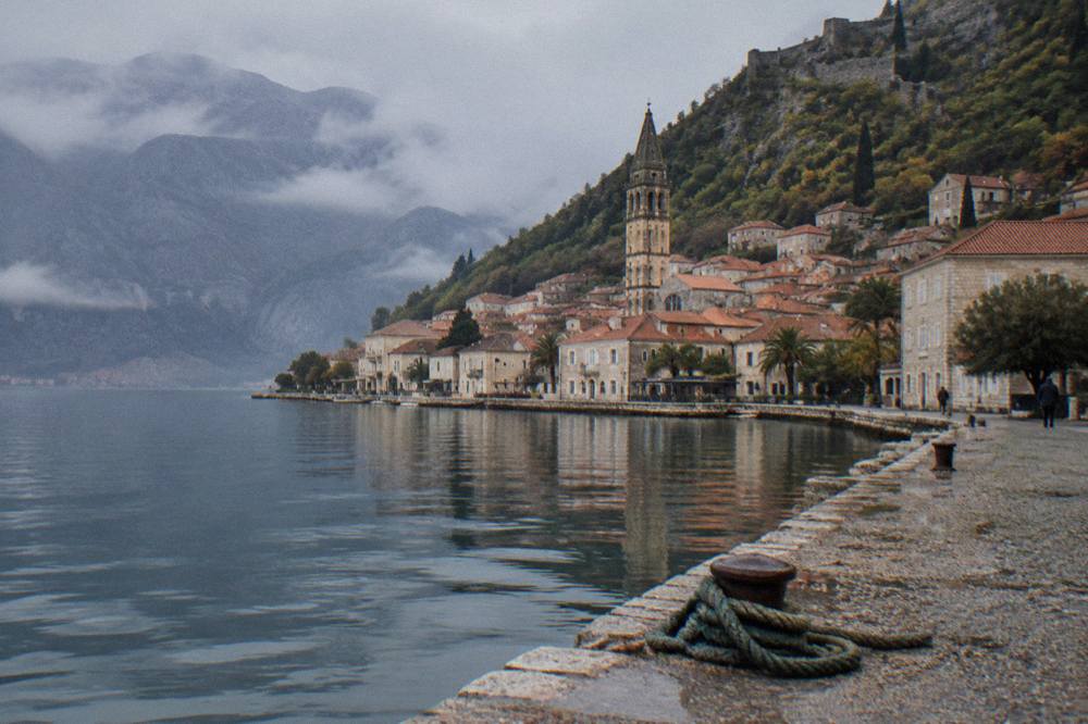 Old stone buildings with terracotta roofs rising up a hillside toward a mountain ridge above a bay.