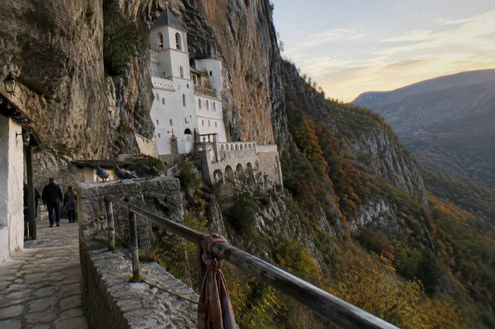 White monastery buildings built into a sheer gray cliff face above a green valley