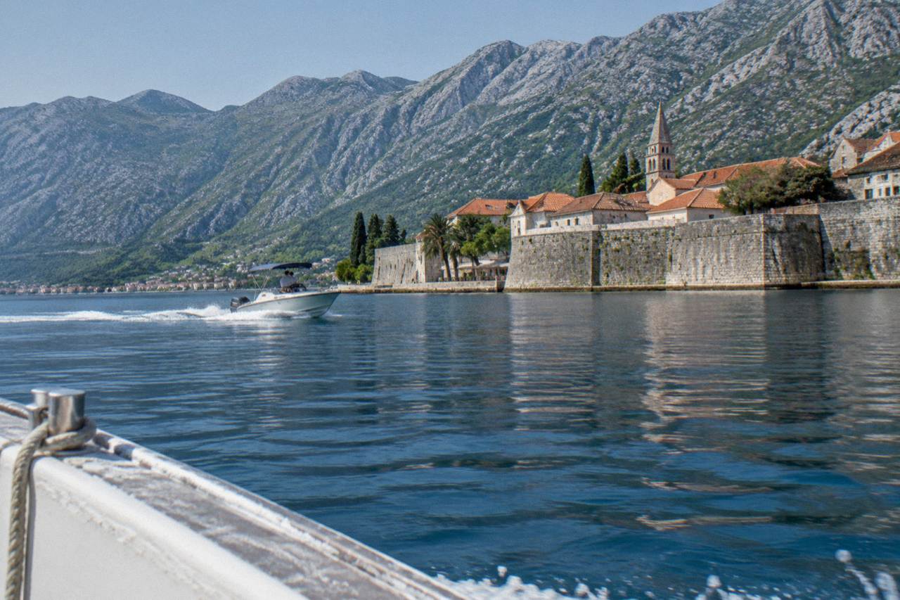 Speedboat on calm blue bay water with steep mountains rising behind a walled old town