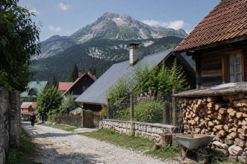 A small mountain town of pitched roofs with pine slopes and grey peaks behind under a clear sky