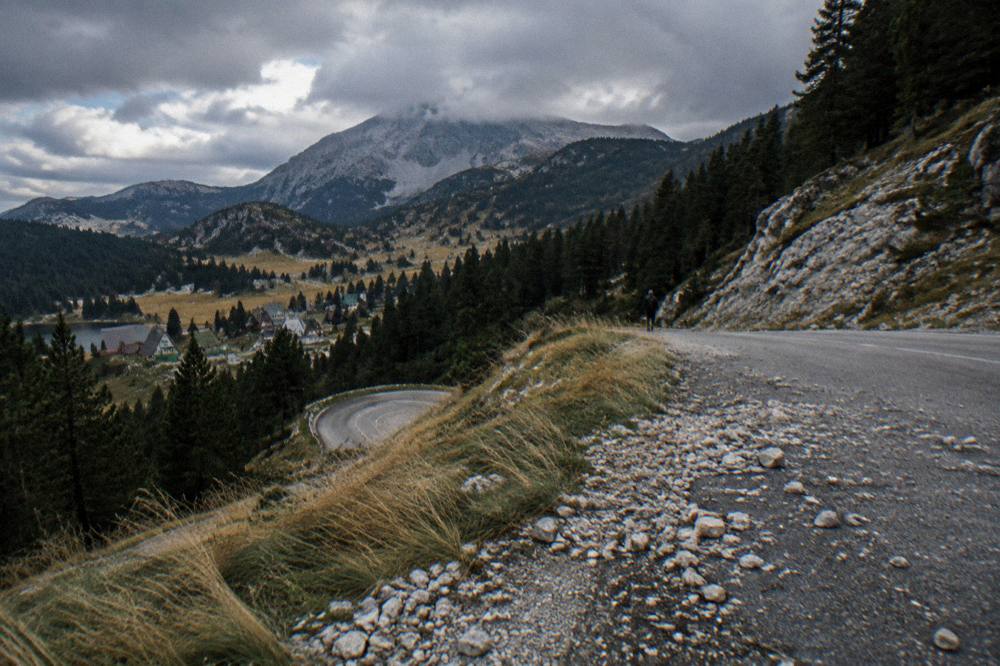 A mountain valley road curving between pine forest and grey limestone peaks under scattered clouds