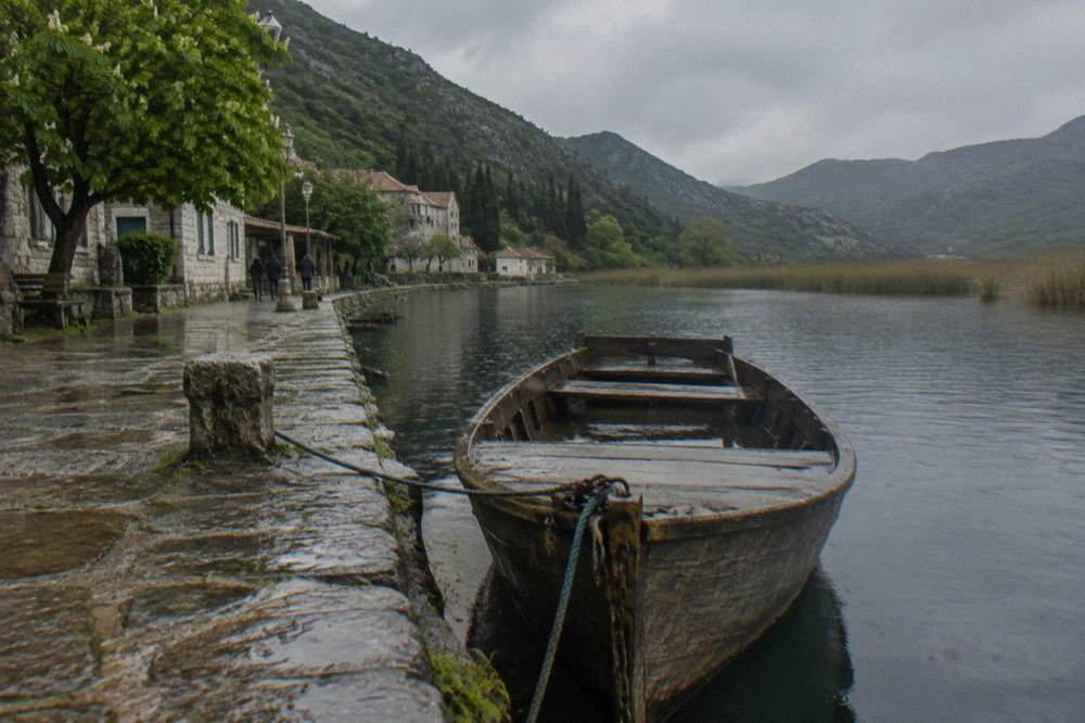 A wooden boat tied at a small stone jetty beside a lakeside village with hills behind