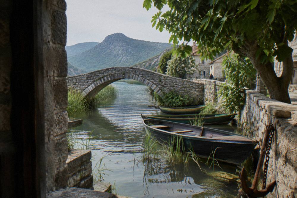 Small stone bridge over a channel leading into a lakeside village with moored wooden boats