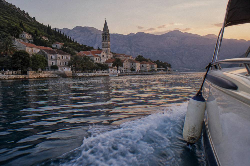 Speedboat passing a stone church tower on the shoreline of the Bay of Kotor with mountains behind