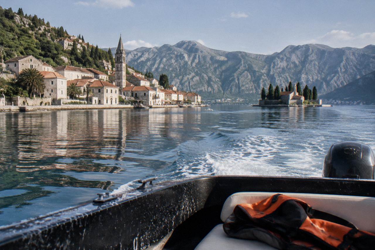 Speedboat crossing calm water toward the stone waterfront of Perast with two small islands in the distance