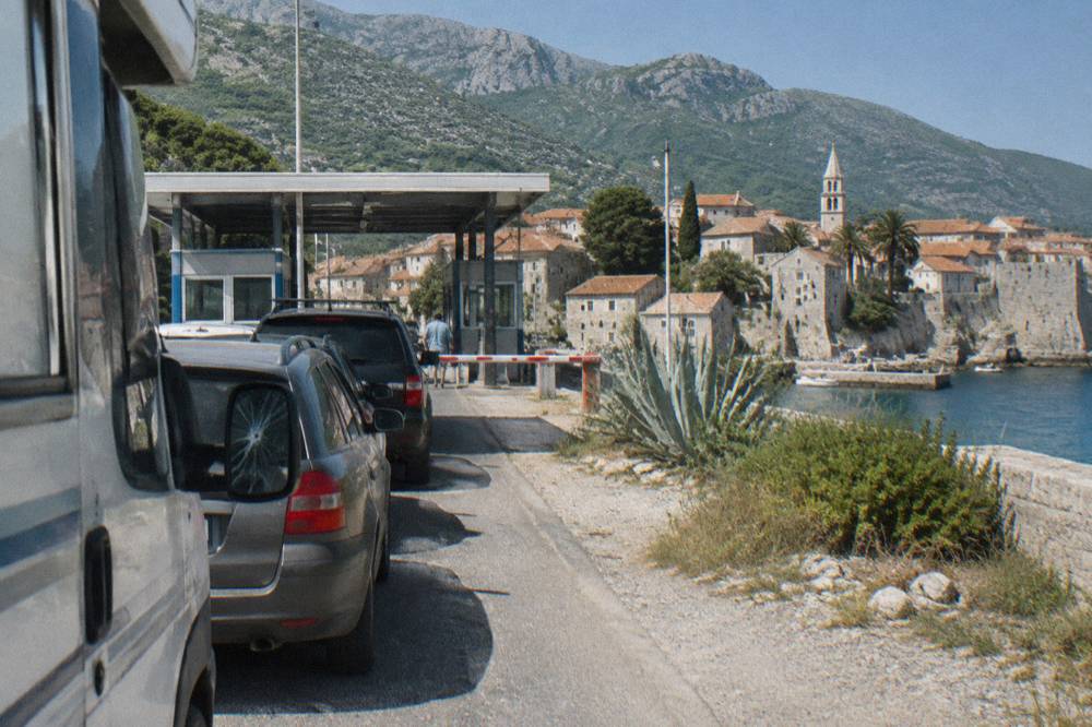 A queue of cars waiting at a striped border barrier under a summer sky