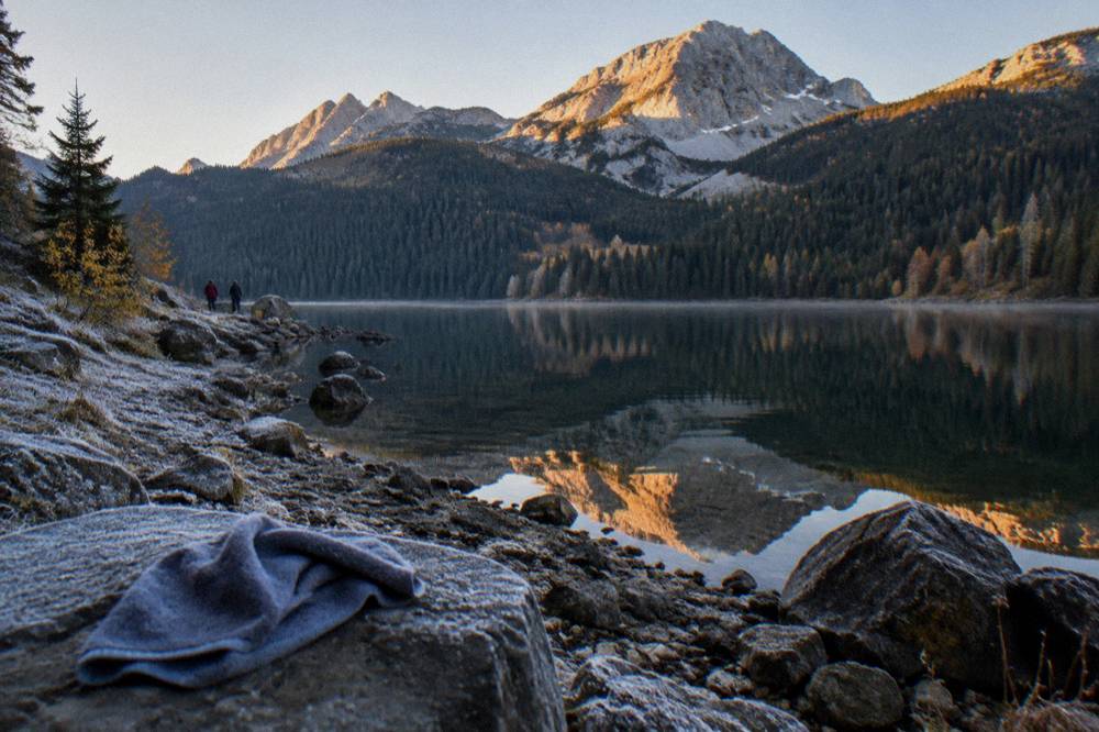 A calm mountain lake reflecting pine forest and a stone shoreline under a clear sky