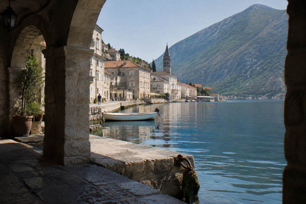 Sunlit shoreline with stone buildings along a calm bay and steep hills behind