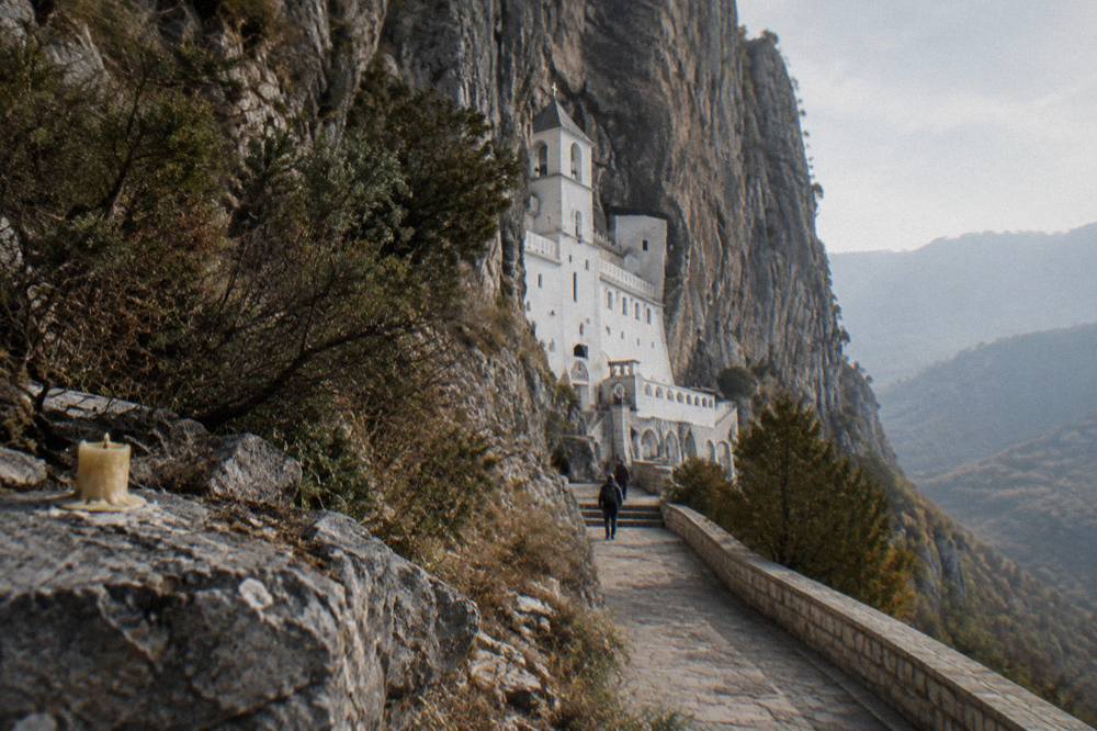 A white monastery church built into the rock of a tall grey cliff