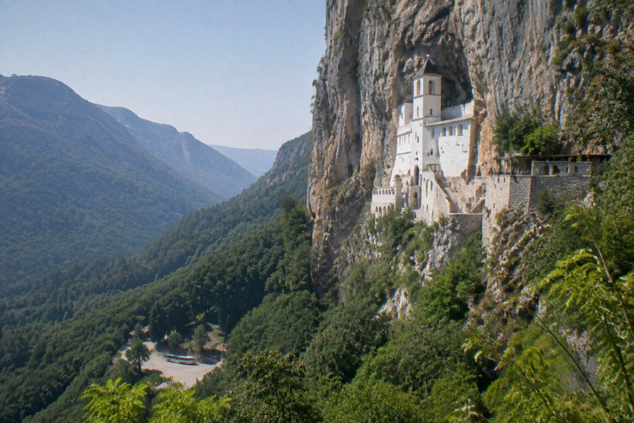 White monastery buildings built into a sheer grey cliff face above forested hills