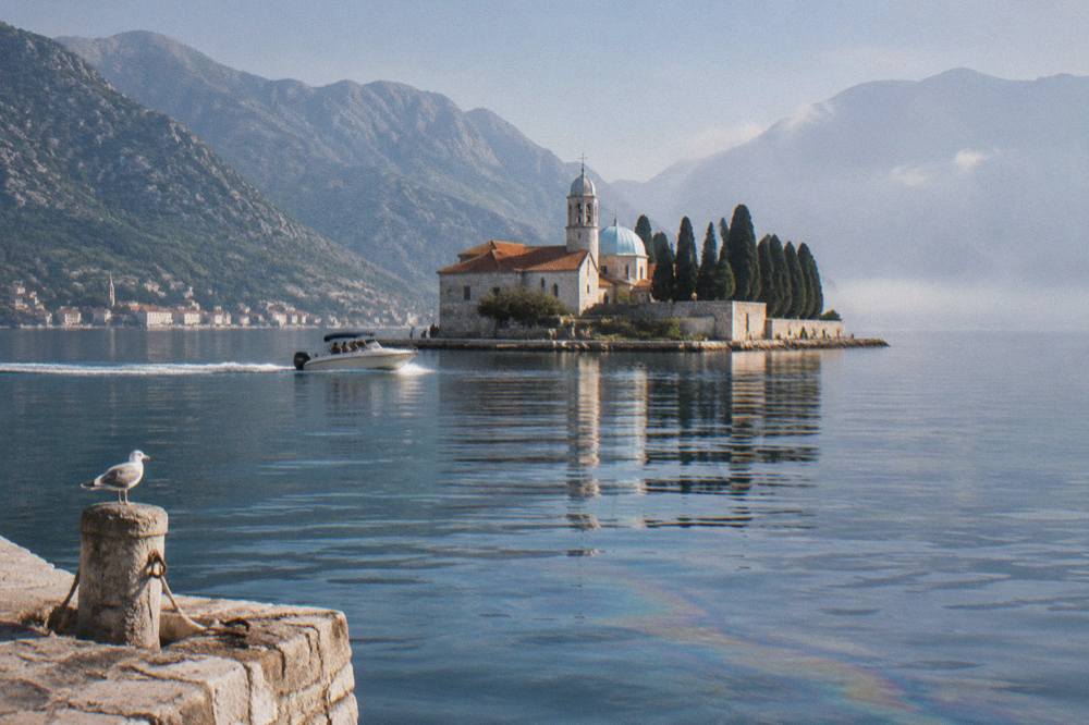 Speedboat on the flat blue water of the Bay of Kotor passing a stone island church