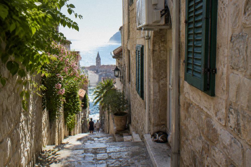 Narrow stone street with shuttered houses leading toward bright sea light in an old coastal town